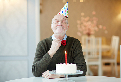 Portrait of happy old man smiling to camera and adjusting bow tie proudly while sitting at table with birthday cake and wearing party hat