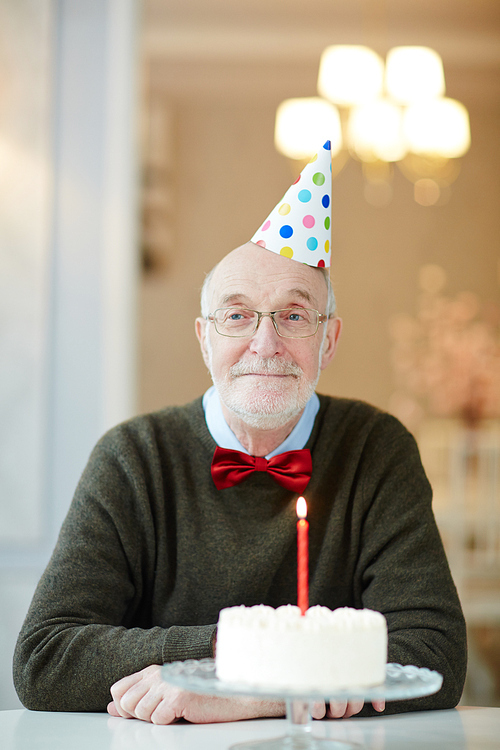 Portrait of nice old man smiling happily to camera while sitting at table with birthday cake and wearing party hat