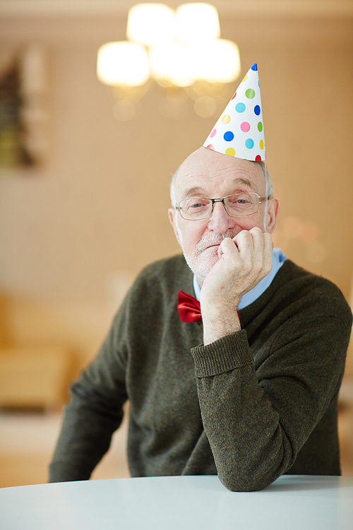 Portrait of nice old man wearing party hat sitting alone at table waiting for guests looking pensive