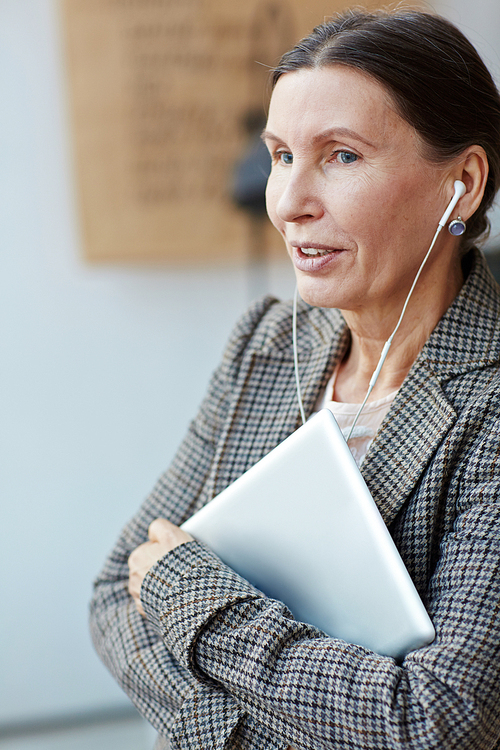Pretty senior woman with laptop in hands looking away dreamily and listening to music with headphones, waist-up portrait