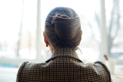 Unrecognizable woman with elegant hair bun wearing houndstooth jacket sitting at panoramic window, back view