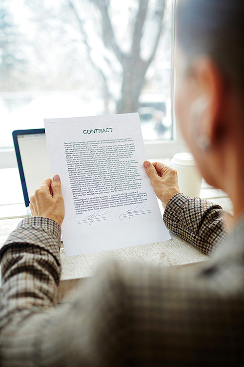 Senior entrepreneur holding signed business contract in hands while sitting at panoramic window of coffeehouse, over shoulder view