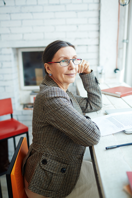 Smiling elegant-looking senior woman wearing eyeglasses sitting in profile and looking out window, waist-up portrait