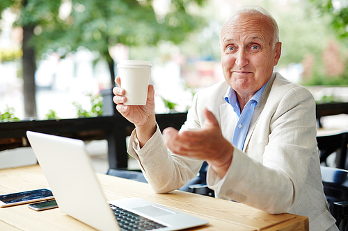 Portrait of enthusiastic elderly man working with digital devices at outdoor coffee shop, smiling happily to camera and enjoying life, gesturing actively