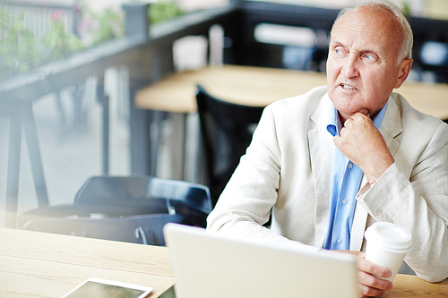 Frowning senior man looking sideways thinking hard while working with laptop at outdoor area of street cafe during coffee break