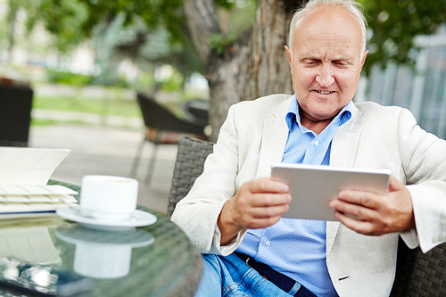 Portrait of elderly man trying to use tablet, looking curious and confused, while relaxing at outdoor seating area of cafe on summer day