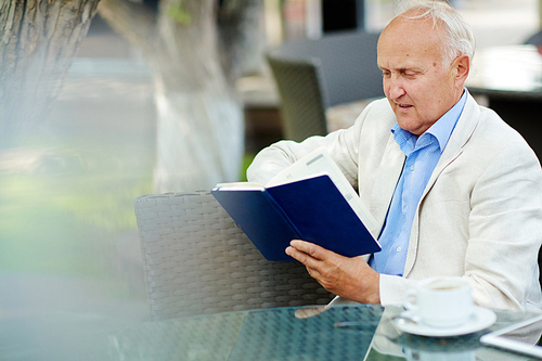 Portrait of elegant mature man looking into book while having coffee break at outdoor area of quiet cafe in shade of tree on summer day