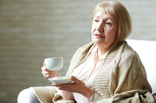 Melancholic senior woman with plaid on shoulders holding coffee mug in one hand and saucer with cookies in other while relaxing in living room