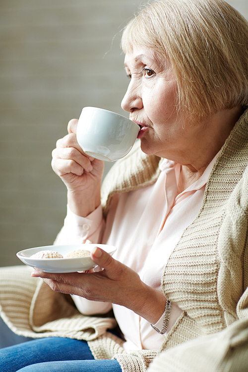 Profile portrait shot of pensive retired woman in jeans and pink shirt drinking coffee with pastry in small cafe