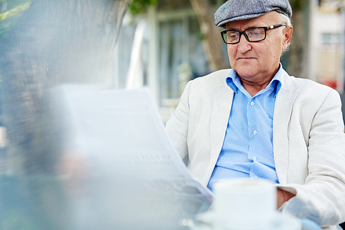 Portrait of pleasant senior man wearing cap and  black rim glasses enjoying his rest in outdoor cafe while reading fresh newspaper in shade of tree on summer day