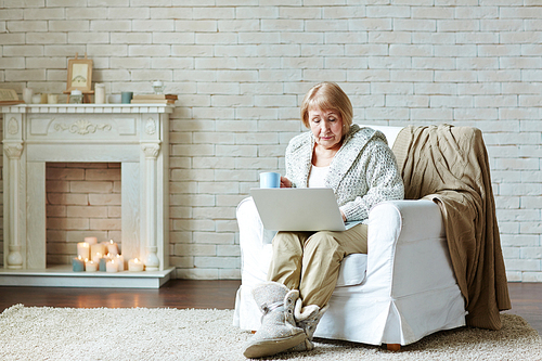 Retired woman in comfortable home clothing ensconced herself in armchair by fireplace and watching her favorite soap opera on laptop in lovely living room