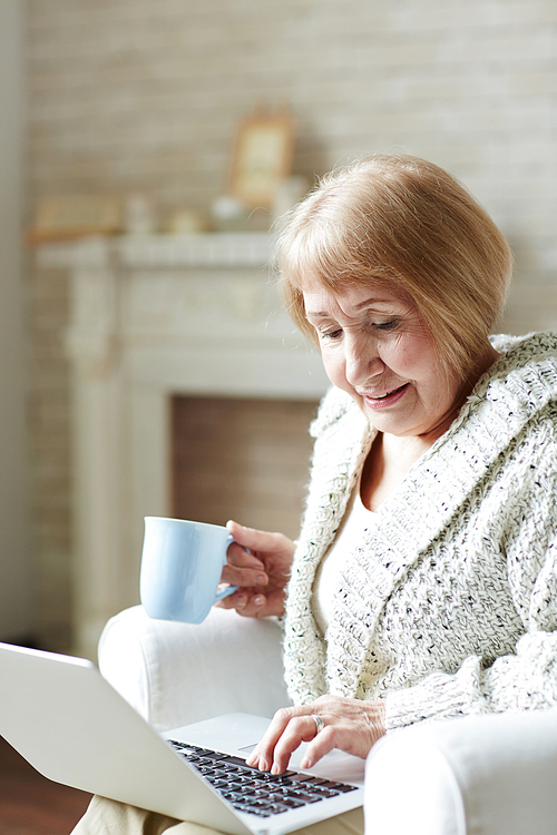 Modern granny sitting at home with cup of coffee and chatting with her granddaughter via social networking site, fireplace decorated with mementos observed behind her