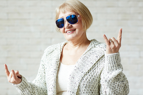 Trendy elderly woman in sunglasses making horn gesture with both hands and  with confident smile against white background