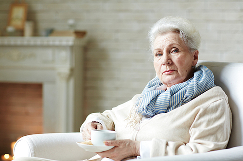 Waist-up portrait of retired woman in striped scarf  with deep blue eyes and drinking tea in living room