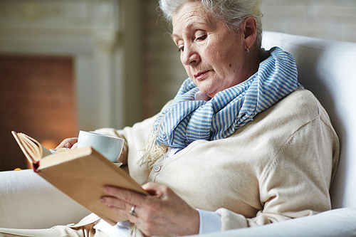 Calm winter evening by fireplace: pretty senior woman sitting back on armchair with interesting book and holding tea mug in hand