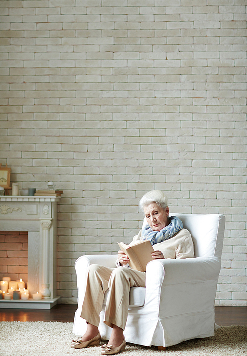 Serious elderly woman wrapped up in reading adventure novel while sitting by fireplace decorated with burning candles, full-length portrait