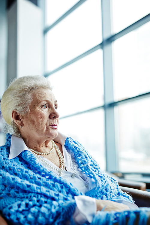 Waist-up portrait of beautiful elderly woman with pearl necklace and cornflower blue knitted shawl sitting on armchair and looking out panoramic window