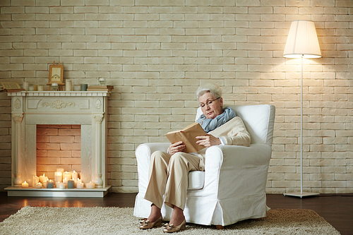 Full-length portrait of elderly woman in eyeglasses focused on reading exiting book while sitting in cozy armchair against white brick wall