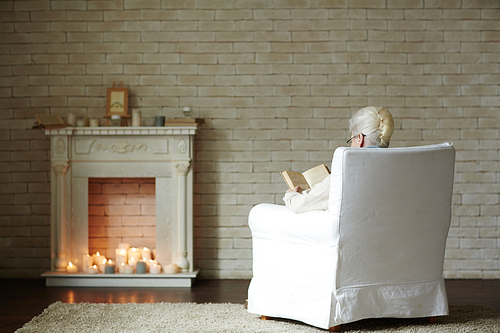 Back view of elderly woman sitting by fireplace and enjoying calm winter evening with interesting book