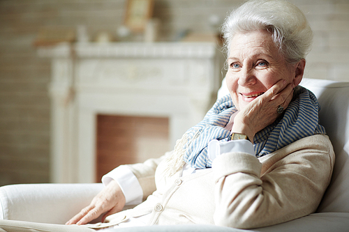 Smiling elderly woman with natural make-up looking away while sitting on cozy armchair by fireplace