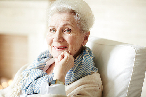 Portrait of beautiful old lady in beige cardigan and striped scarf sitting in armchair,  and smiling cheerfully