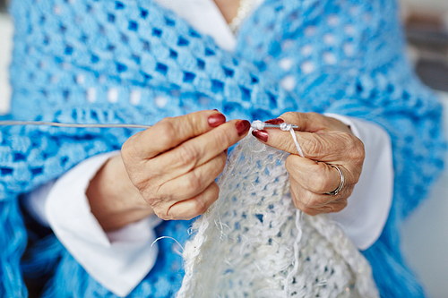 Close-up shot of wrinkled hands of senior woman holding unfinished knitted scarf and crochet needle in hands