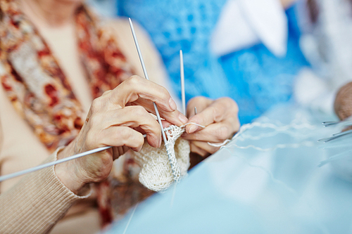 Close-up shot of elderly woman knitting white socks for her little granddaughter with help of three needles, blurred background