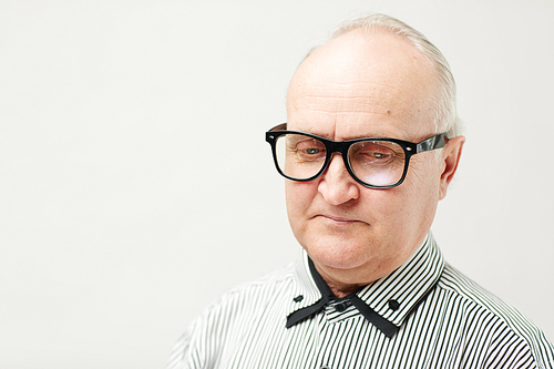 Close-up portrait of upset elderly man in eyeglasses and striped shirt looking down thoughtfully on white wall background