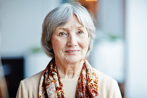 Head and shoulders portrait of pensive elderly woman in beige sweater and colorful kerchief on shoulders looking away while standing at window