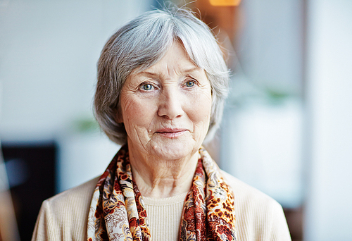 Portrait of beautiful grey-haired senior woman standing against blurred background and looking away thoughtfully
