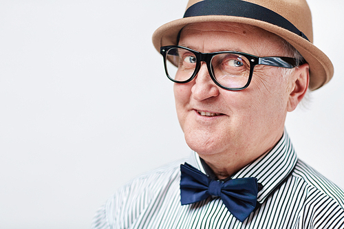 Close-up portrait of charming senior man in glasses, beige hat and striped shirt with bowtie looking to the side and smiling playfully