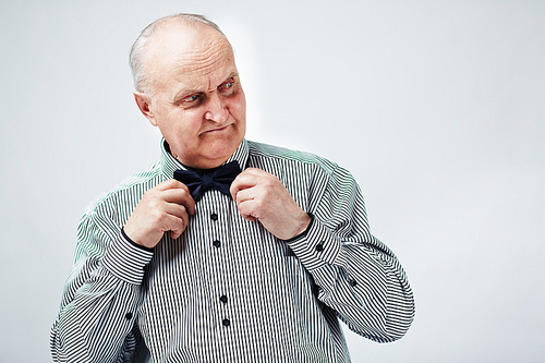 Waist-up portrait of elegant elderly man in striped shirt fixing his bow tie and looking to the side pensively