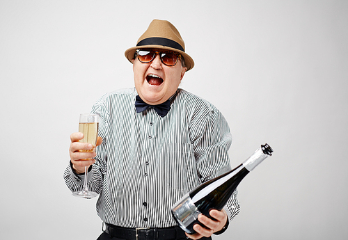 Waist-up portrait of dressed-up retired man holding a bottle of champagne and laughing cheerfully with his mouth wide open