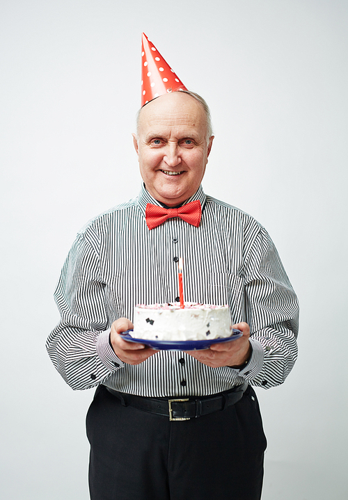 Portrait of charming old man in party hat and bow tie standing straight, holding birthday cake with one candle and smiling cheerfully