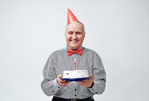 Portrait of happy stylish old man in party hat holding birthday cake with one candle and  with bright toothy smile