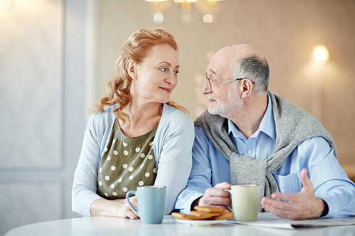 Amorous mature woman looking at her husband while listening to him by cup of tea