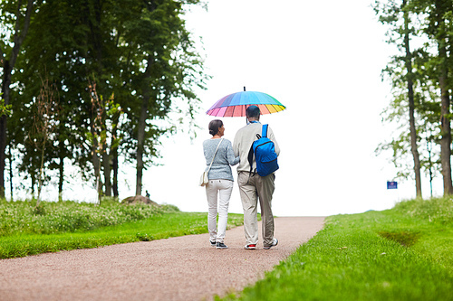 Aged retired couple walking down country road under rainbow-colored umbrella