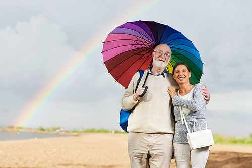 Happy aged couple under colorful umbrella walking against cloudy sky and rainbow