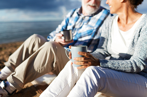 Senior couple with mugs relaxing on sandy beach at weekend