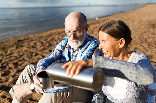 Restful seniors sitting on seashore and having hot tea from thermos