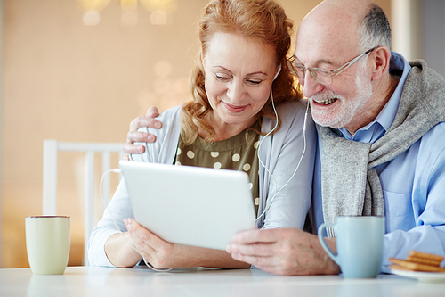 Portrait of smiling elderly couple using tablet watching videos with earphones looking at screen and embracing home at table