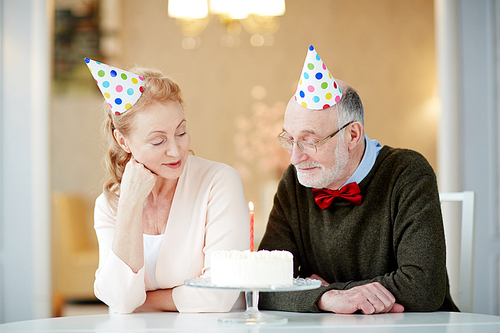 Married couple sitting by table and looking at burning candle in birthday cake