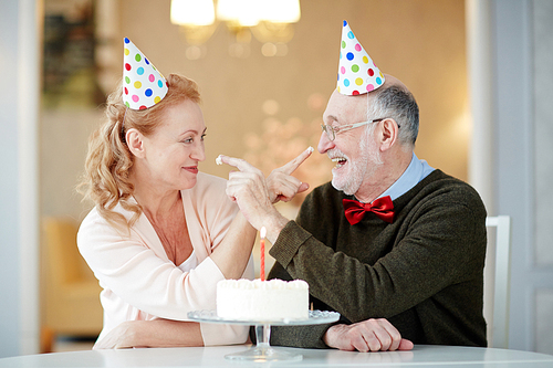 Portrait of playful senior couple having fun celebrating birthday together sitting at table with cake and wearing party hats trying to smudge each other with cream