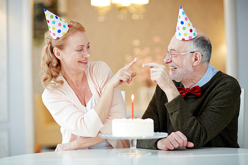 Portrait of playful senior couple laughing and  having fun celebrating birthday together sitting at table with cake and wearing party hats trying to smudge each other with cream