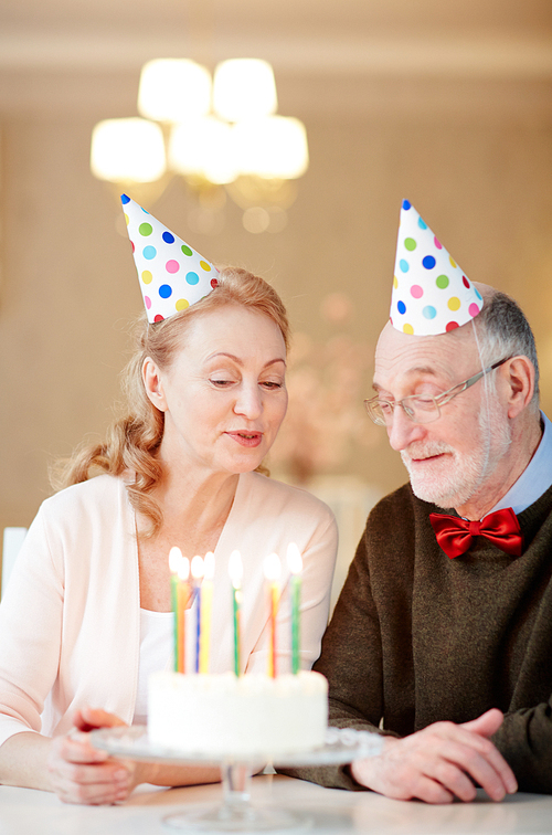 Happy mature man and his wife in birthday caps looking at burning candles in cake