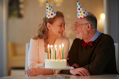 Portrait of loving senior couple celebrating birthday together sitting at table with cake and wearing party hats looking tenderly in each others eyes