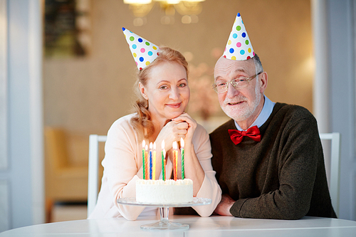 Portrait of loving senior couple celebrating birthday together sitting at table with cake and wearing party hats smiling and  