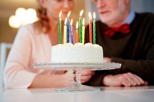 Closeup shot of small creamy birthday cake with candles on stand and nice senior couple sitting at table in background
