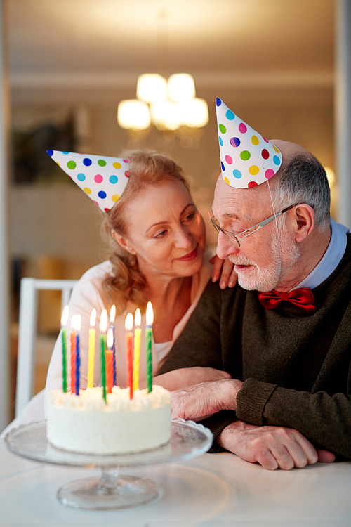 Portrait of loving senior couple celebrating birthday together embracing,  sitting at table with cake and wearing party hats, old man going to blow the candles and make a wish