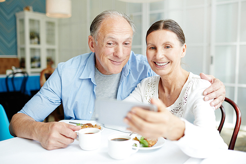 Smiling senior couple making selfie by table in restaurant during dessert